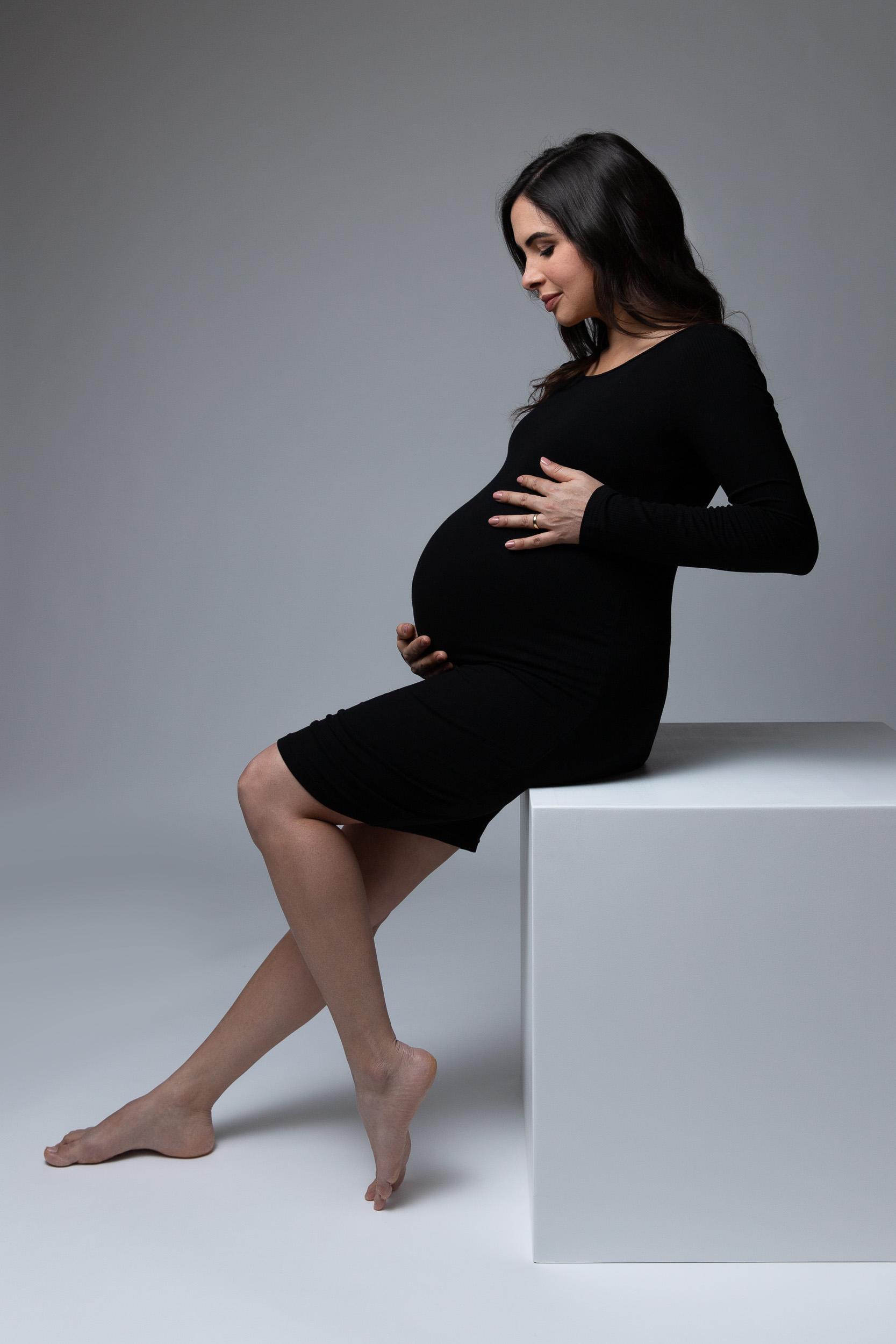 Maternity portrait in a fitted black dress, seated in profile on a white block against a soft grey backdrop
