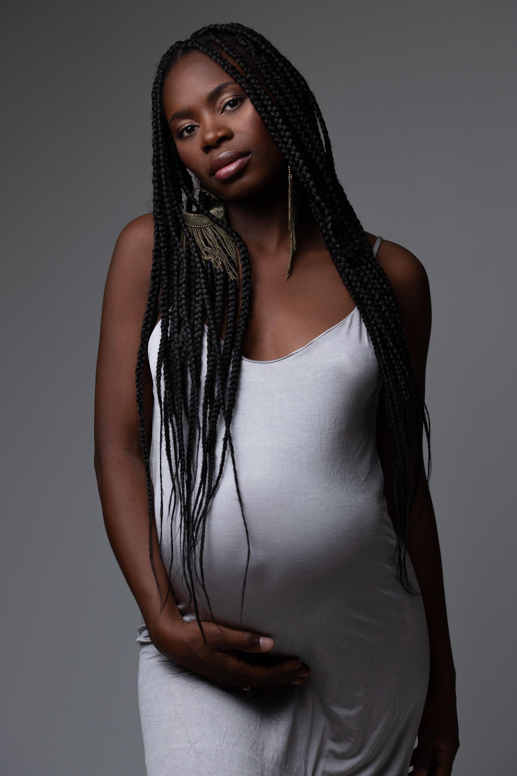 Maternity portrait with long braids in a white slip dress, hand resting on the belly, against a grey studio backdrop
