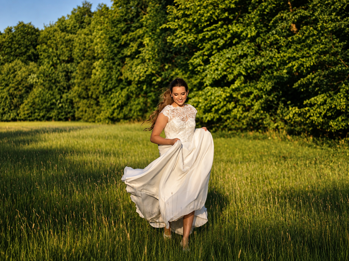Bride smiling and walking through a sunlit grassy field, wedding dress caught mid-movement