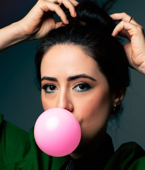 Playful editorial portrait blowing a pink bubblegum bubble, teal graphic eyeliner, green wardrobe