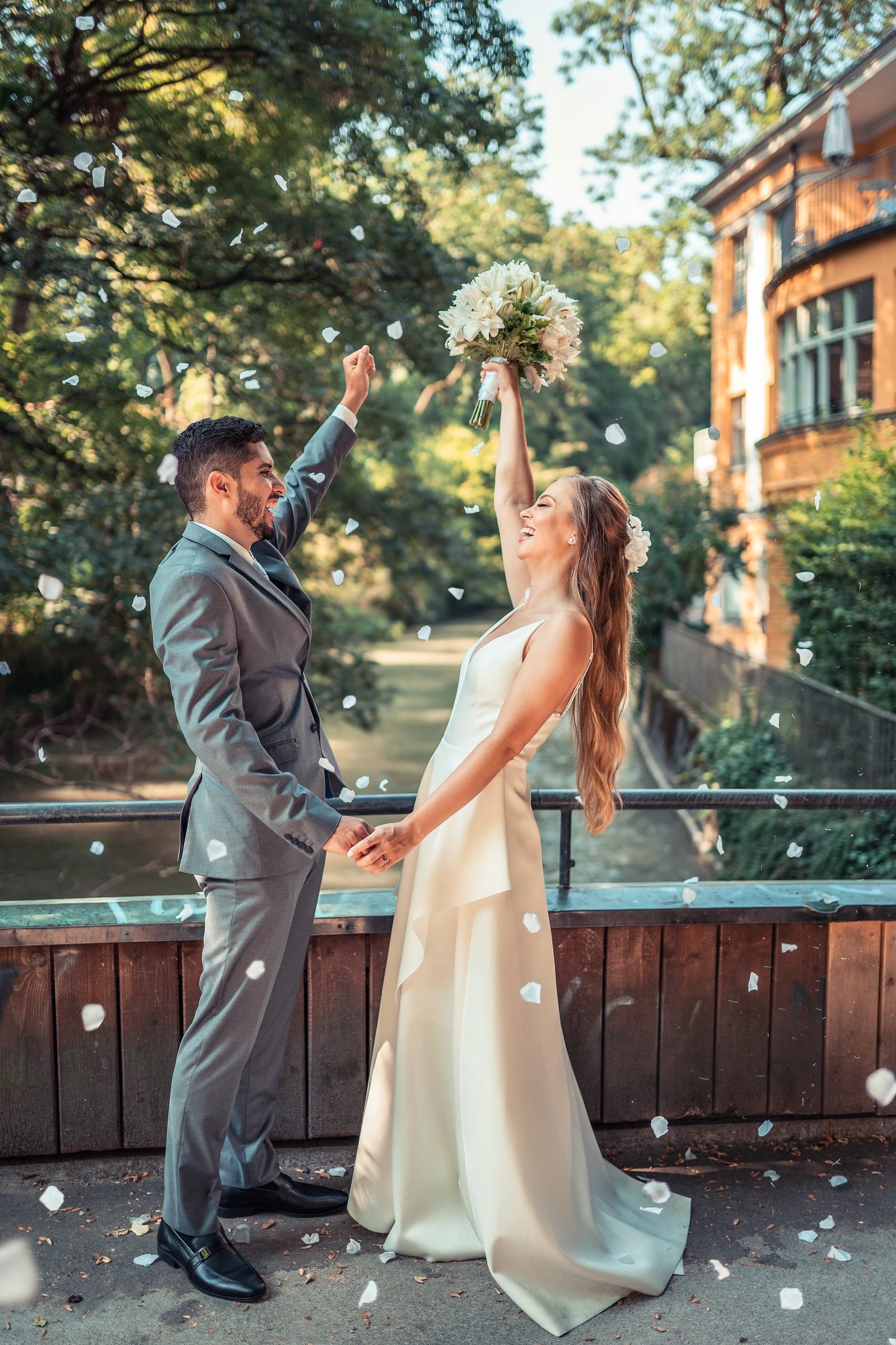Bride and groom celebrating under falling petals, bride raising her bouquet, long hair worn loose