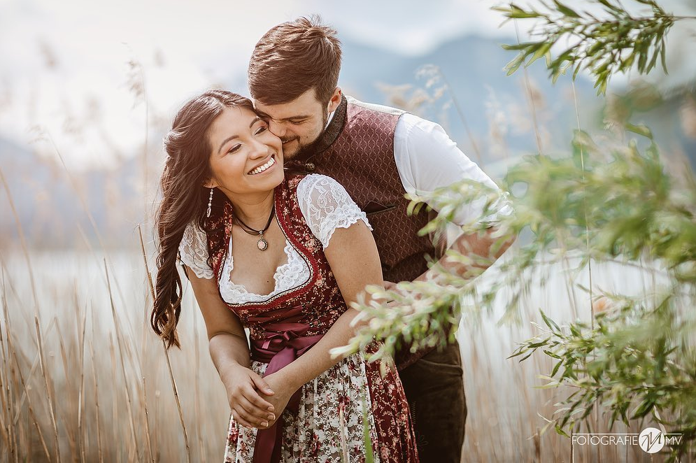 Couple in traditional Bavarian dirndl and tracht embracing by a lake with mountains behind