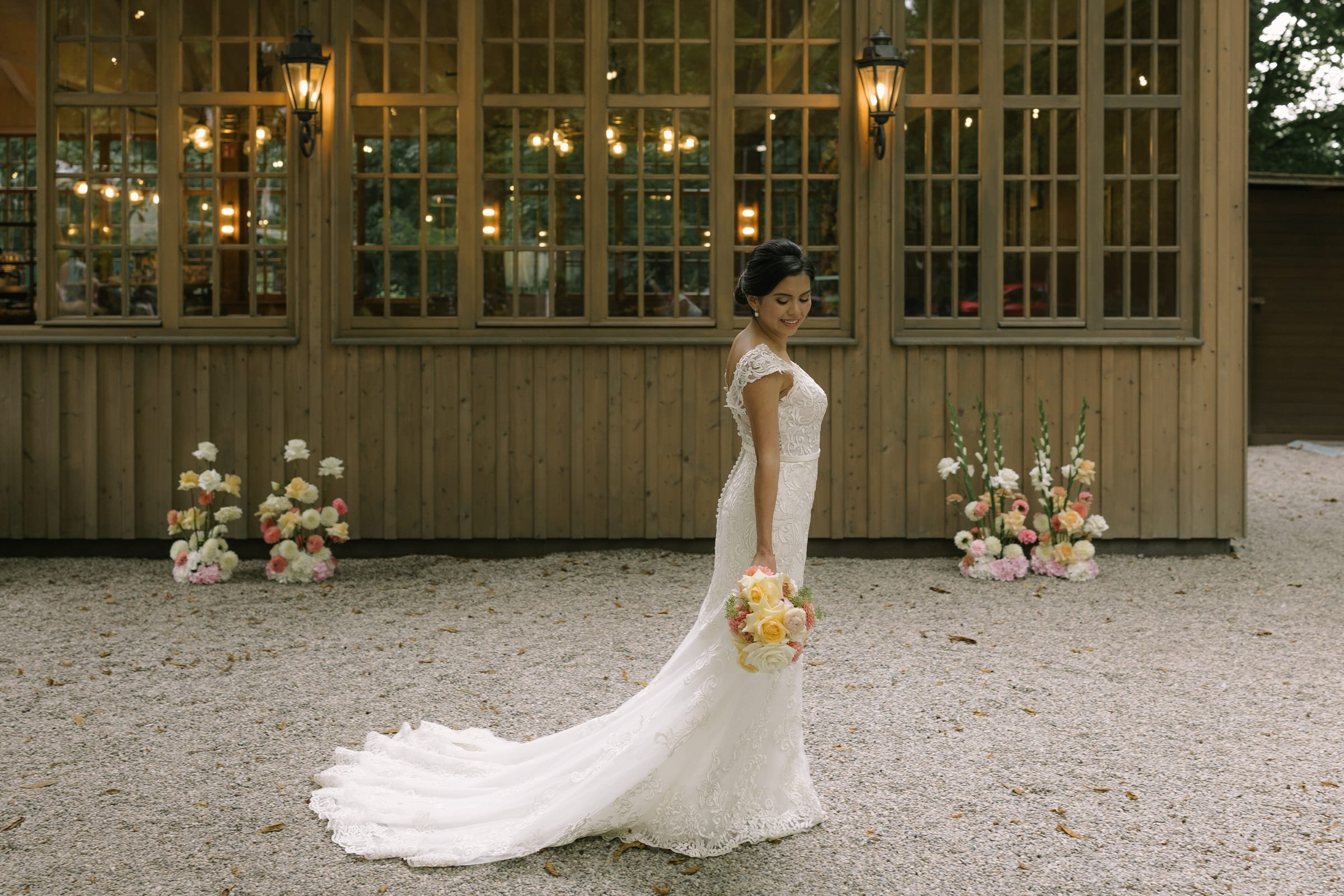 Bride in a lace gown with a long train in front of a glass-fronted pavilion decorated with flowers