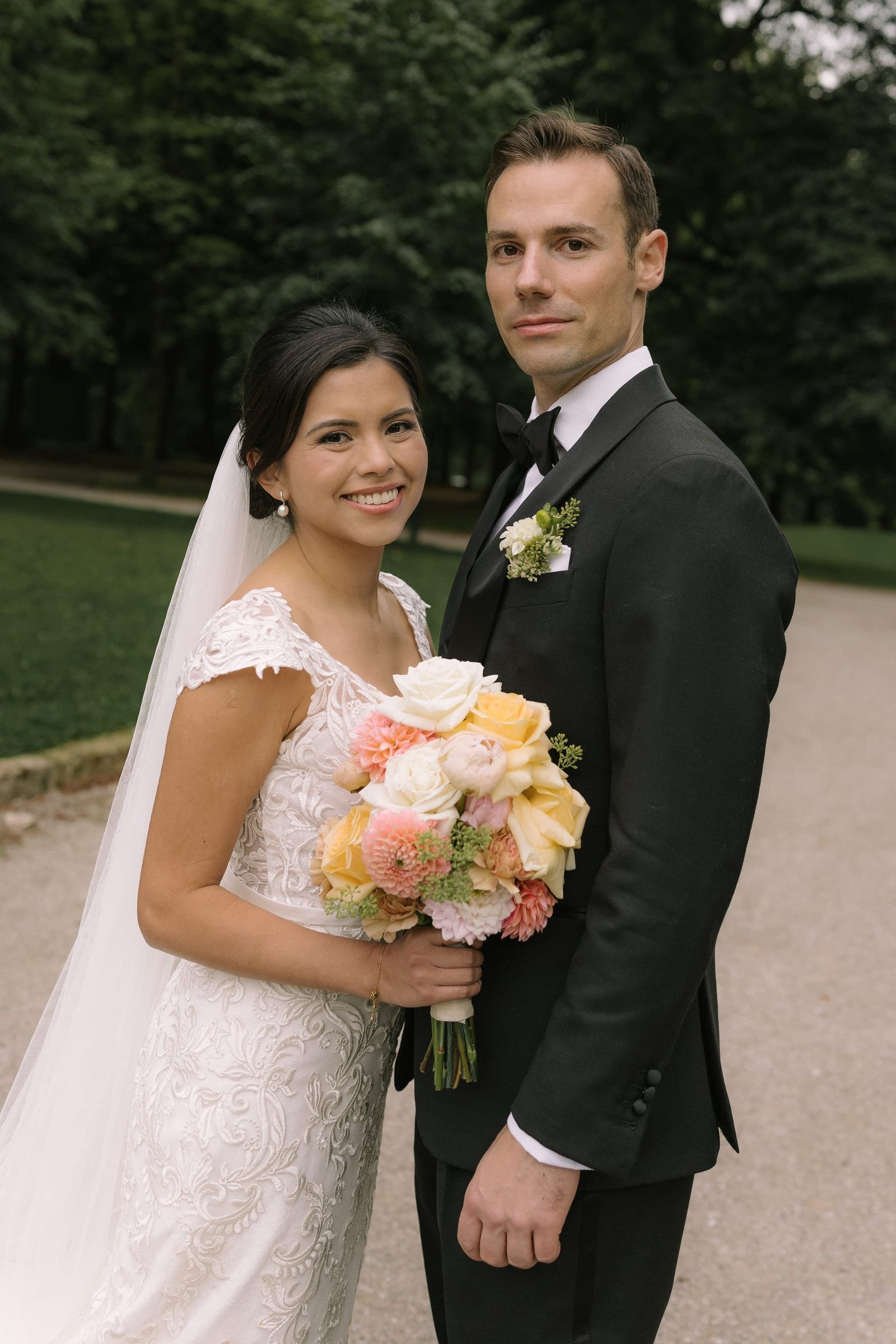 Bride with a veil and pastel bouquet beside the groom in a park, polished bridal makeup