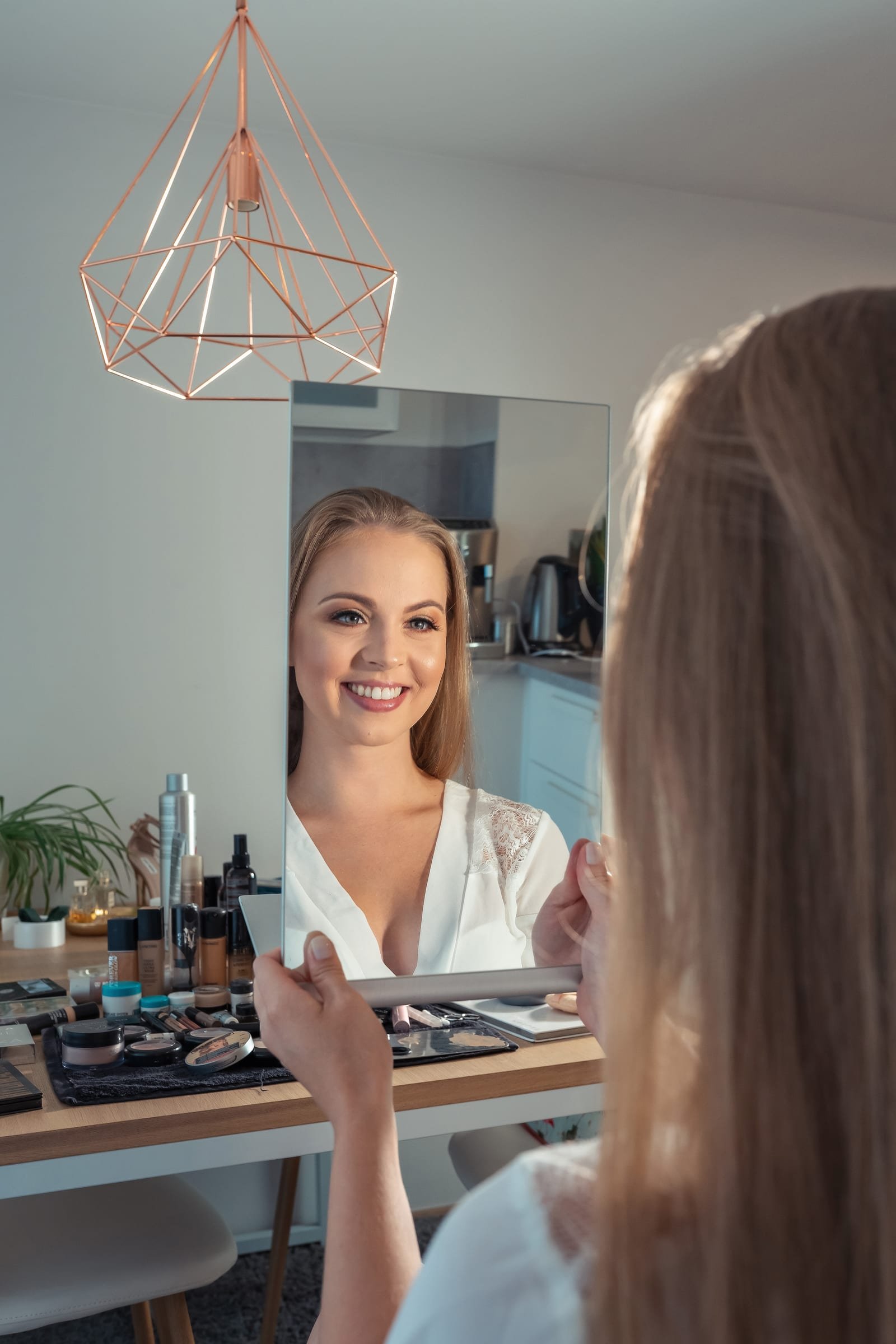 Bride smiling into a mirror during her makeup session, products laid out on the table