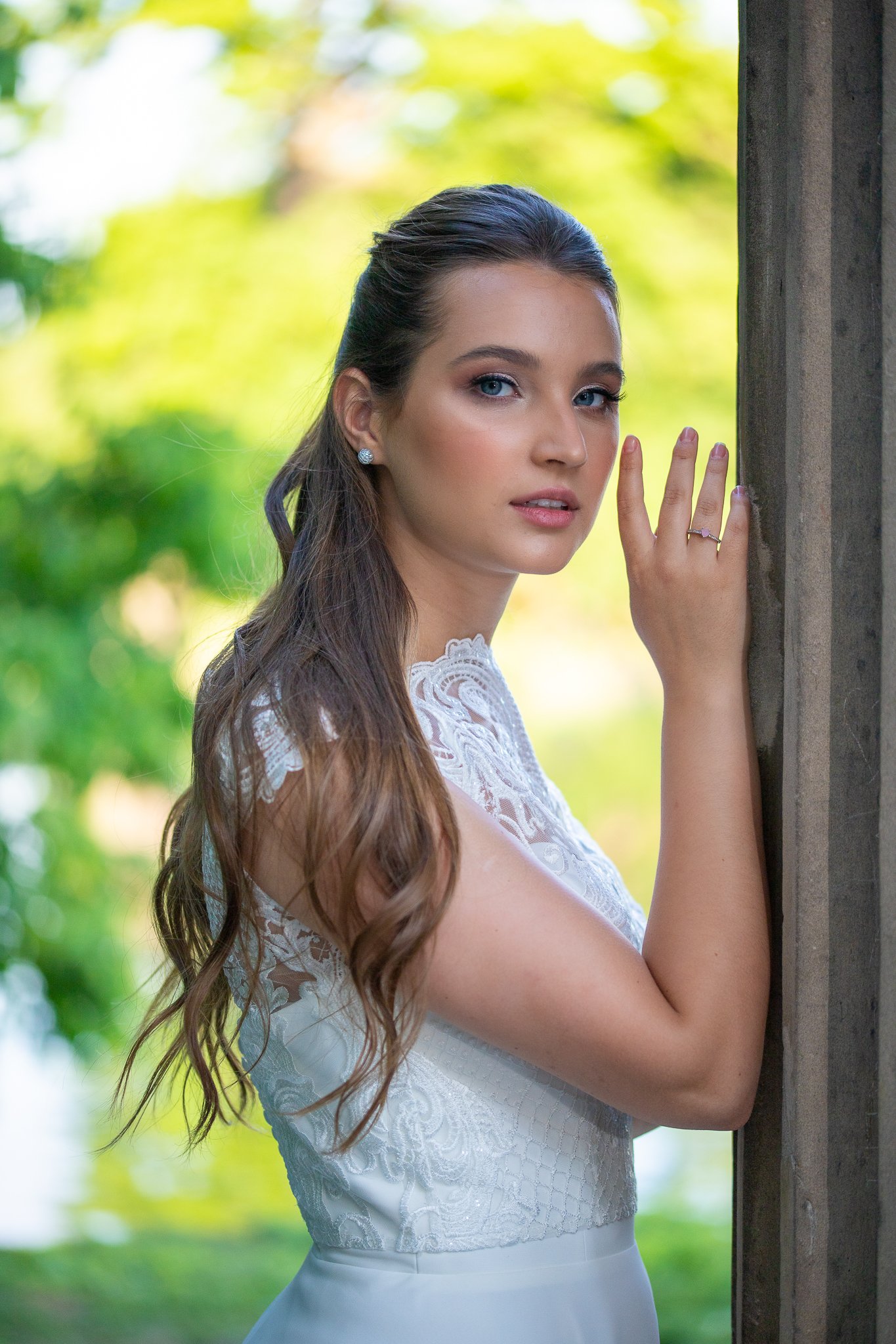 Bride with a sleek half-up hairstyle, long waves and luminous makeup, leaning against a wooden post