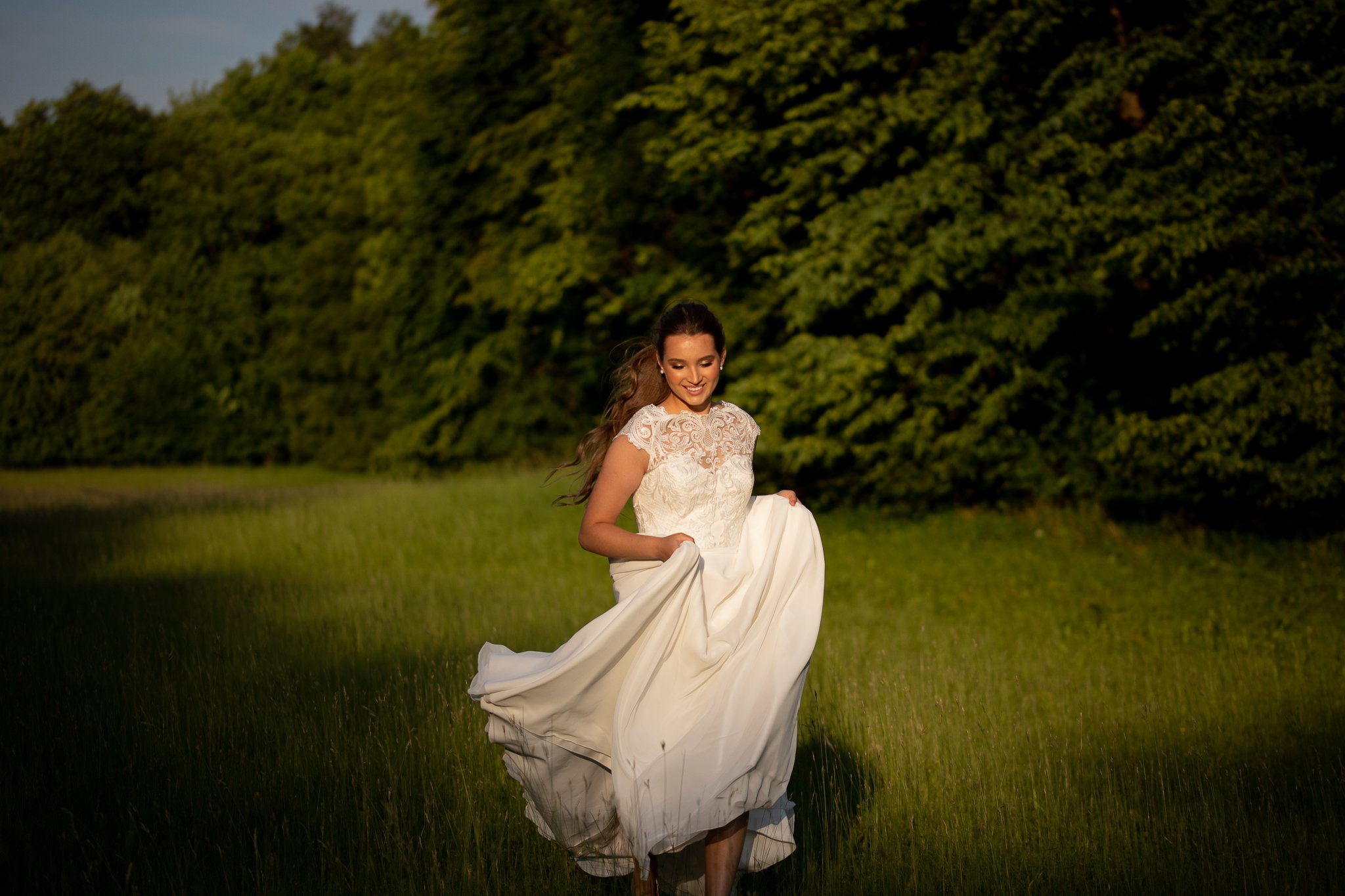 Bride in a lace wedding dress lifting her skirt in a sunlit meadow, natural bridal makeup and loose hair