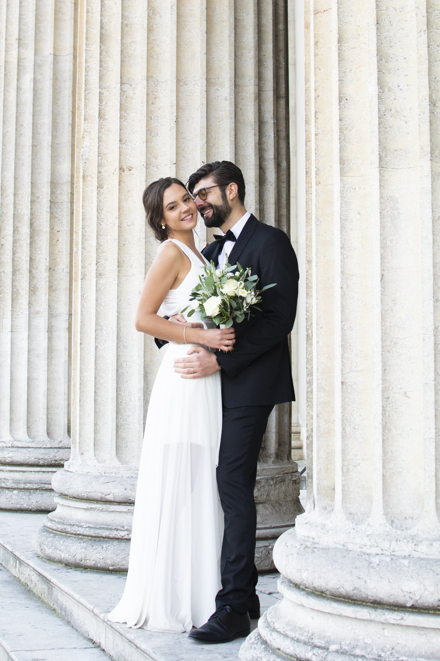 Bride and groom embracing between stone columns, bride holding a white bouquet