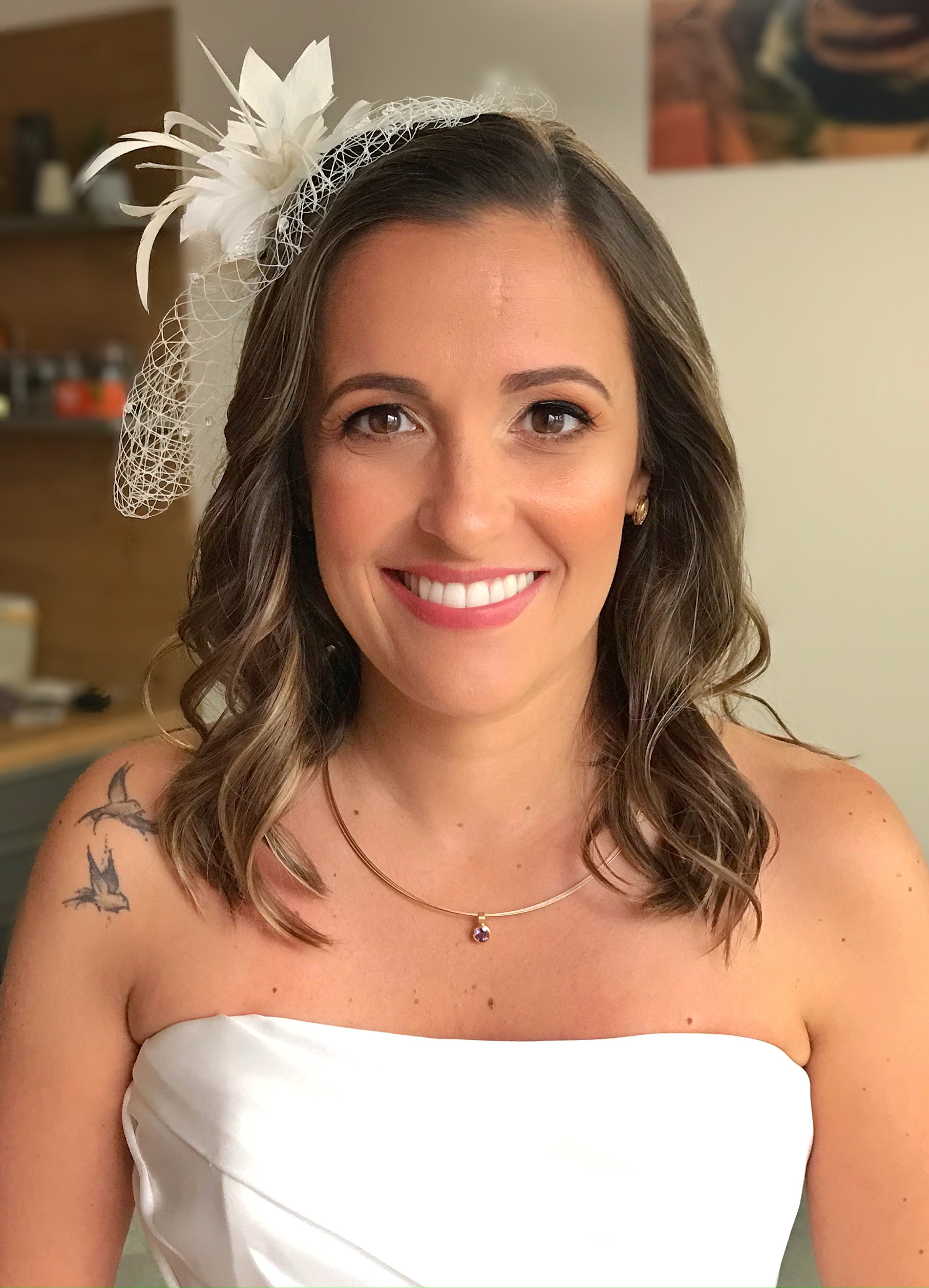 Smiling bride with shoulder-length waves, a feather fascinator and soft bridal makeup in a strapless dress